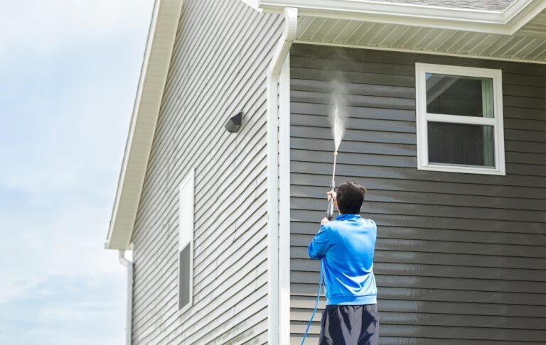 Man in blue jacket clean dusk and dirt from exterior siding and under roof by high-pressure nozzle spray with water soap cleaner