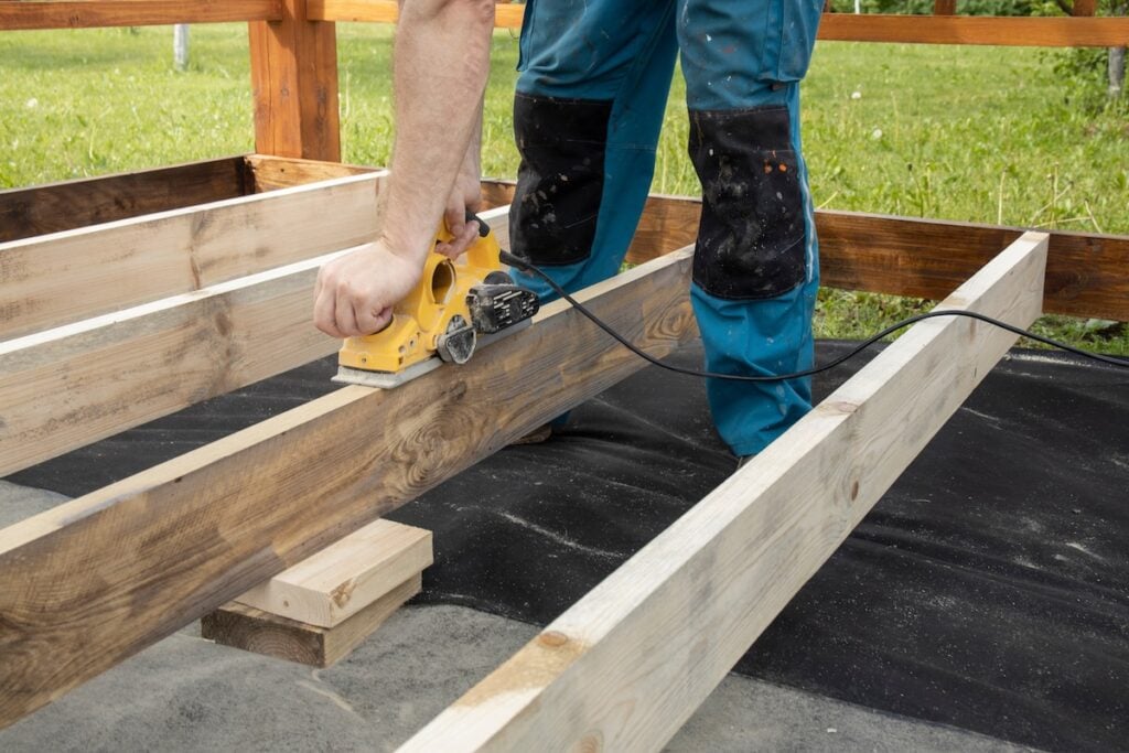 A carpenter wearing blue pants sands a piece of wood using a power planer. The wood is part of a deck frame being built.