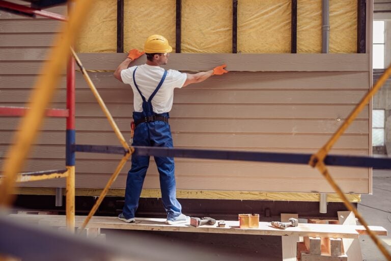 Back view of young man builder wearing safety helmet and work overalls while installing exterior siding